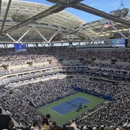 Tennis court with a blue surface is surrounded by a large stadium filled with spectators. The stadium has a massive metal roof structure supported by tall beams. An American flag flies in the distance.