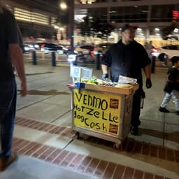 A person in a black shirt and gloves stands next to a yellow and brown cart on a brick-lined sidewalk at night. The cart displays food items and a sign advertising "Venmo Hotzelle HogCosh". Other people are partially visible in the background, along with parked cars and city lights.