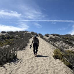 A person walks on a sandy path between dunes covered with sparse vegetation. The path slopes upwards towards a horizon with a blue sky and scattered clouds.