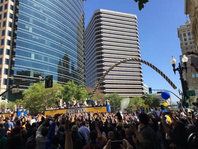 A championship parade celebration with crowds of fans gathered around buses carrying players through downtown city streets, with confetti streamers and balloons visible against a backdrop of modern office buildings.