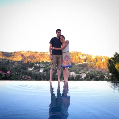 A couple embraces while standing at the edge of an infinity pool, with rolling hills dotted with houses and golden sunset light visible in the background.