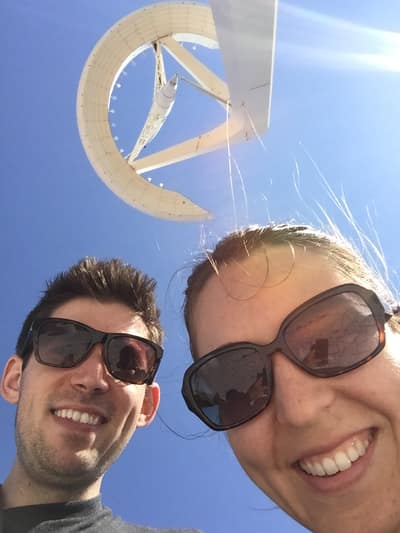 Two people wearing sunglasses take a selfie in front of a large white sculptural sundial against a bright blue sky.