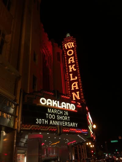 The iconic Fox Oakland Theater marquee glows with red neon lights against the night sky, advertising "Too Short 30th Anniversary" for March 26. The historic theater's ornate vertical sign displays "FOX OAKLAND" in bright yellow letters surrounded by decorative red bulbs.