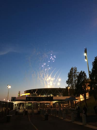Fireworks explode in the twilight sky above a modern stadium with distinctive angular architecture, while outdoor dining areas and walkways are illuminated below.