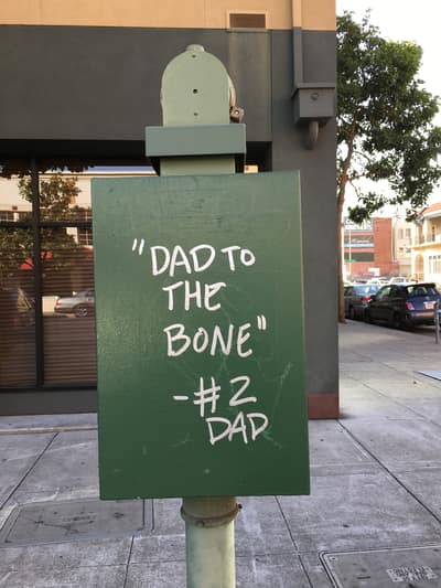A green street sign with white text reading "DAD TO THE BONE" and "#2 DAD" stands on a sidewalk in an urban area with buildings and parked cars in the background.