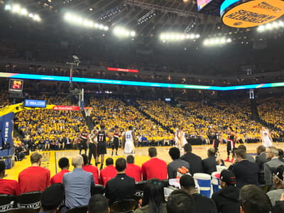 Basketball players compete on the court at Oracle Arena during an NBA playoff game, with a packed crowd of fans wearing yellow filling the stands behind them.