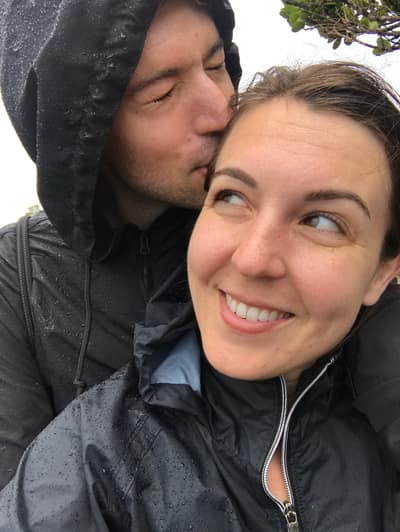A couple embraces outdoors in the rain, with the man kissing the woman's forehead while she smiles broadly at the camera. Both are wearing dark rain jackets with water droplets visible on the fabric.