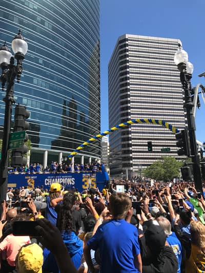 A crowd of fans gathers around a blue parade bus celebrating the Golden State Warriors' championship victory, with tall glass office buildings forming the urban backdrop in downtown.