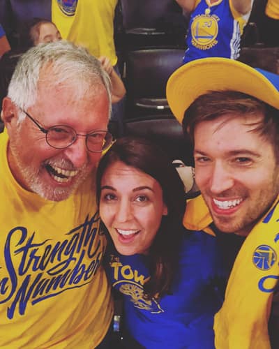 Three smiling people pose for a selfie at a Golden State Warriors basketball game, wearing team colors of blue and yellow while seated in arena stands filled with other fans.