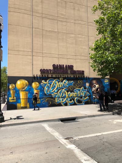 A large mural reading "Dubs Dynasty" decorates the exterior wall of the Oakland Convention Center, featuring golden championship trophies and a player portrait in blue and gold colors celebrating the Golden State Warriors.
