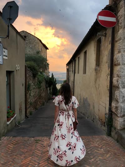 A woman in a white floral dress walks down a narrow cobblestone street in a historic European town during golden hour, with weathered stone buildings lining both sides and a dramatic sunset sky visible in the distance.