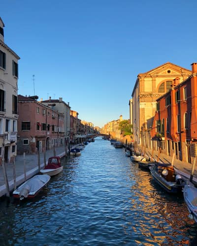 A picturesque Venetian canal lined with colorful historic buildings in warm sunset light, with small boats moored along both sides and clear blue water reflecting the golden architecture.