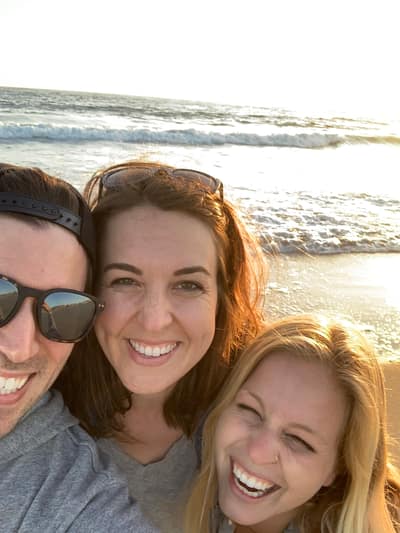 Three people smile together at the beach during golden hour, with ocean waves and sunlight visible in the background.