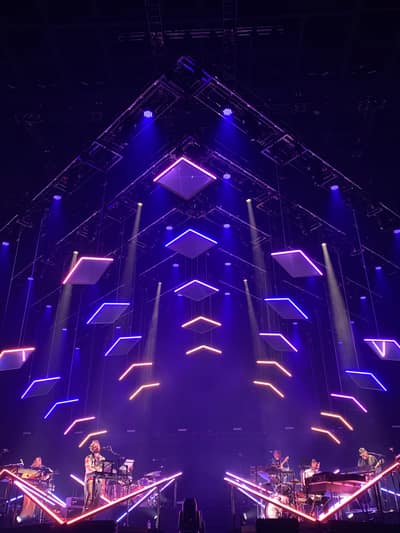 A live music performance takes place on stage beneath a dramatic geometric light installation featuring suspended illuminated diamond shapes and angular LED strips in purple, blue, and orange hues.