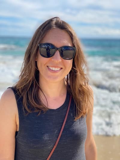 A smiling woman with long brown hair and sunglasses stands on a beach, wearing a dark sleeveless top with ocean waves and blue sky visible in the background.