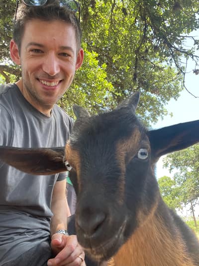 A smiling man in a gray t-shirt poses for a selfie with a brown and black goat under leafy green trees on a sunny day.