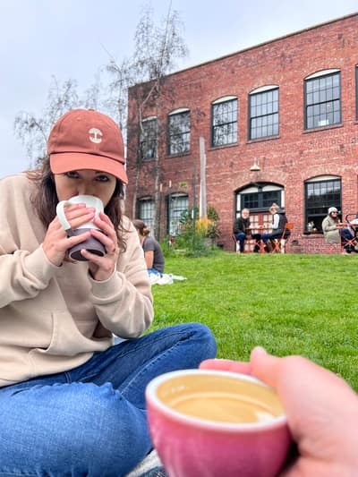 A woman in a rust-colored baseball cap and beige sweater sits on grass drinking from a white mug, with someone holding a pink coffee cup in the foreground and a red brick building with arched windows in the background.