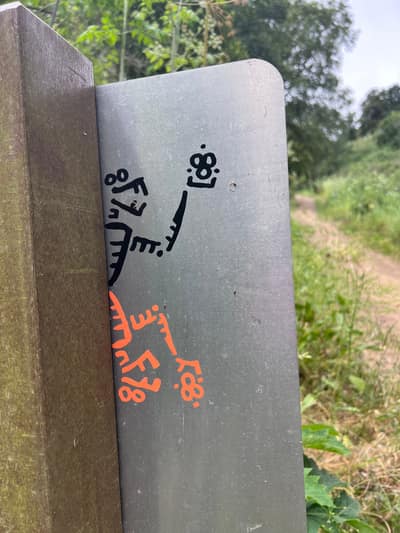 A metal signpost displays black and orange graffiti or stickers in an outdoor setting with lush green trees and vegetation in the background.