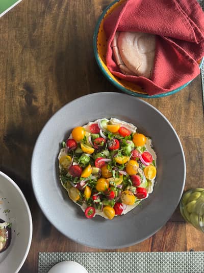 A colorful salad with cherry tomatoes, fresh herbs, and other vegetables is served on a gray plate at a wooden table, with tortillas nearby.