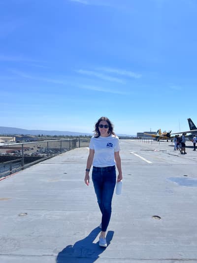 A woman in a white t-shirt and jeans walks along an airport runway or tarmac, with aircraft visible in the background under a clear blue sky.