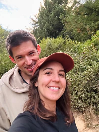 A smiling couple takes a selfie outdoors, with the woman wearing a brown baseball cap in front and the man in a light-colored hoodie behind her, surrounded by lush green trees and vegetation.