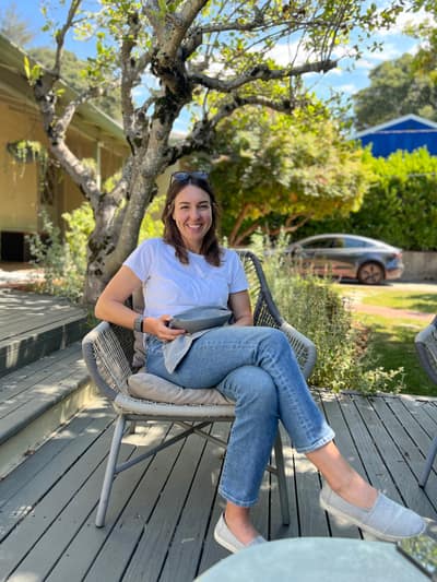 A woman in a white t-shirt and jeans sits smiling in a wicker chair on a wooden deck, surrounded by trees and residential buildings on a sunny day.