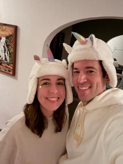 Two people wearing matching white unicorn hats with ears and horns smile at the camera while standing in a home interior with white walls and an arched doorway.
