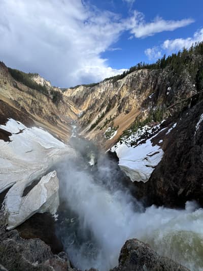 A powerful waterfall cascades down through a dramatic rocky canyon with snow-covered ledges and forested slopes under a blue sky with white clouds.