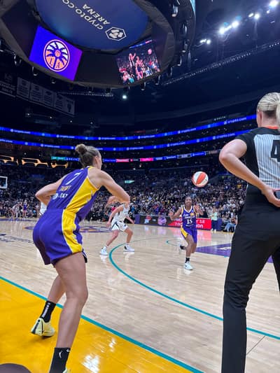 A WNBA basketball game in progress at a packed arena, with players in purple and gold uniforms on the court and a referee in striped shirt observing the action.