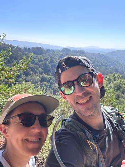 Two hikers wearing sunglasses and caps smile at the camera while taking a selfie on a mountain trail, with rolling forested hills and mountains visible in the background under a clear blue sky.