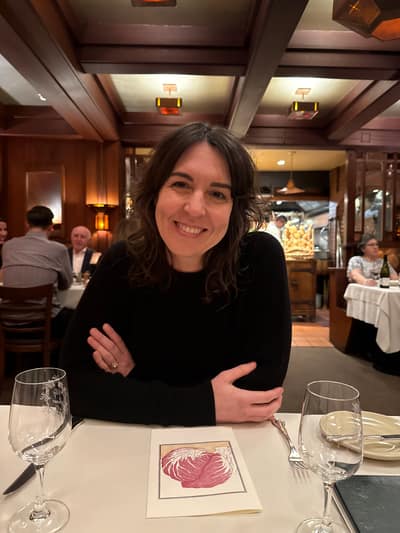 A woman in a black sweater sits smiling at an upscale restaurant table with wine glasses and a menu, with her arms crossed in a relaxed pose. The restaurant features elegant wood paneling, coffered ceilings, and warm lighting typical of a fine dining establishment.