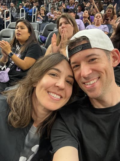 A smiling couple takes a selfie together at what appears to be a sporting event, with crowds of spectators visible in the stadium seating behind them.