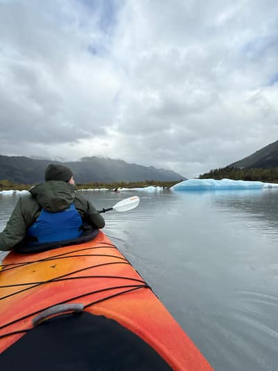 A kayaker in an orange kayak paddles through calm waters dotted with blue icebergs, with snow-capped mountains visible under a cloudy sky in the background.