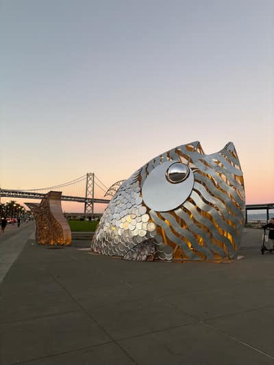 A large metallic fish sculpture with reflective scales and a circular eye sits on a waterfront plaza at sunset, with the Bay Bridge visible in the background.