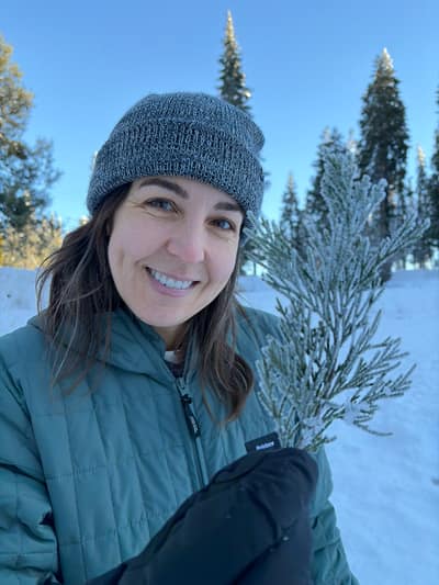 A smiling woman in winter clothing and a knit beanie holds a snow-covered evergreen branch while standing in a snowy landscape with trees and blue sky in the background.