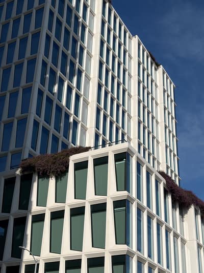A modern white office building with a geometric glass facade features terraced levels with green vegetation growing from planters on the lower floors, photographed against a clear blue sky.