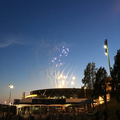 Fireworks explode in the twilight sky above a modern stadium with