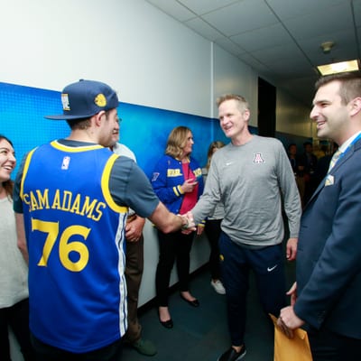 A man in a blue NBA jersey with "SAM ADAMS 76" shakes hands with another
