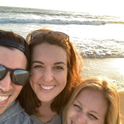 Three people smile together at the beach during golden hour, with ocean