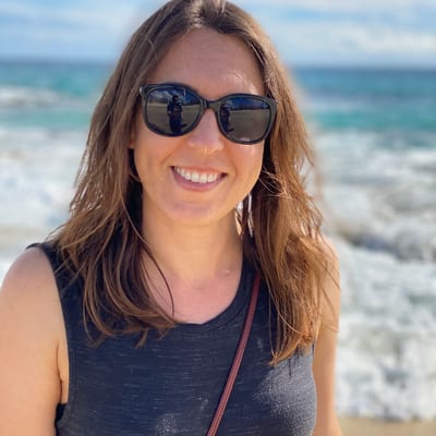 A smiling woman with long brown hair and sunglasses stands on a beach,