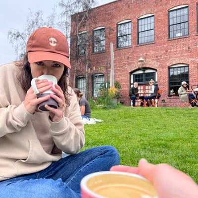 A woman in a rust-colored baseball cap and beige sweater sits on grass