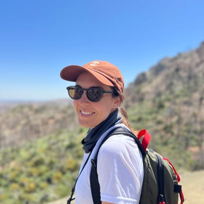 A smiling woman wearing sunglasses, an orange baseball cap, and a backpack