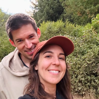 A smiling couple takes a selfie outdoors, with the woman wearing a brown