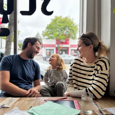 A happy family of three sits at a cafe table by a large window, with