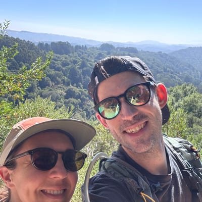 Two hikers wearing sunglasses and caps smile at the camera while taking a