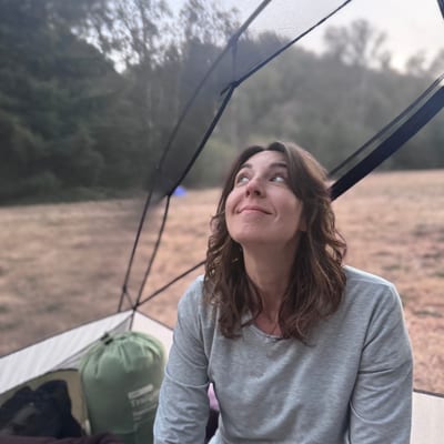 A woman in a gray long-sleeved shirt sits inside a tent, looking up with a