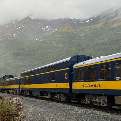 A blue and yellow Alaska Railroad passenger train travels through a misty