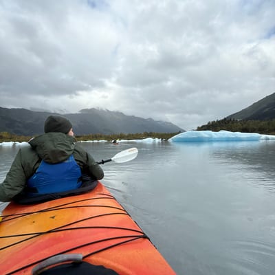 A kayaker in an orange kayak paddles through calm waters dotted with blue