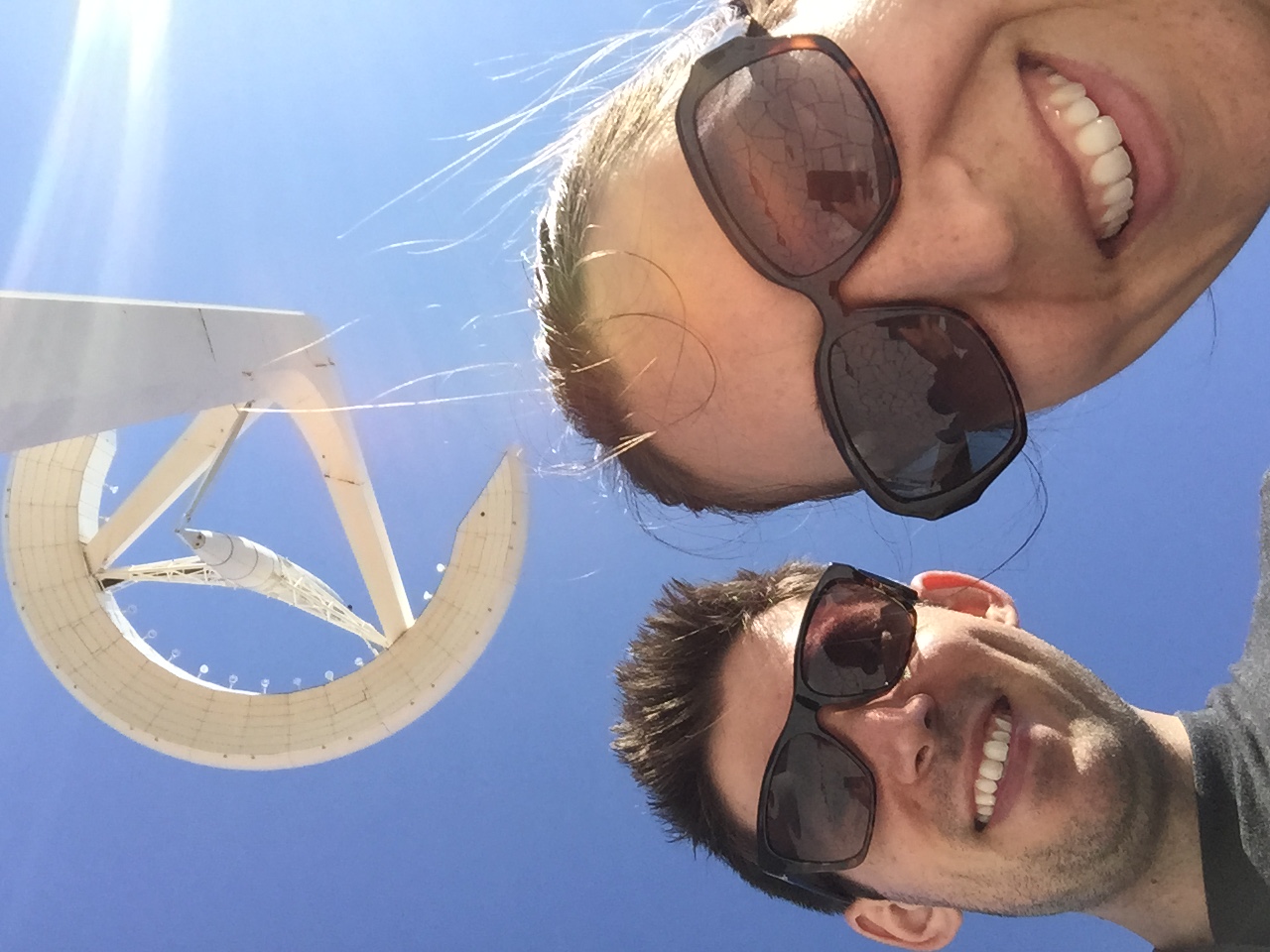 Two people wearing sunglasses take a selfie in front of a large white