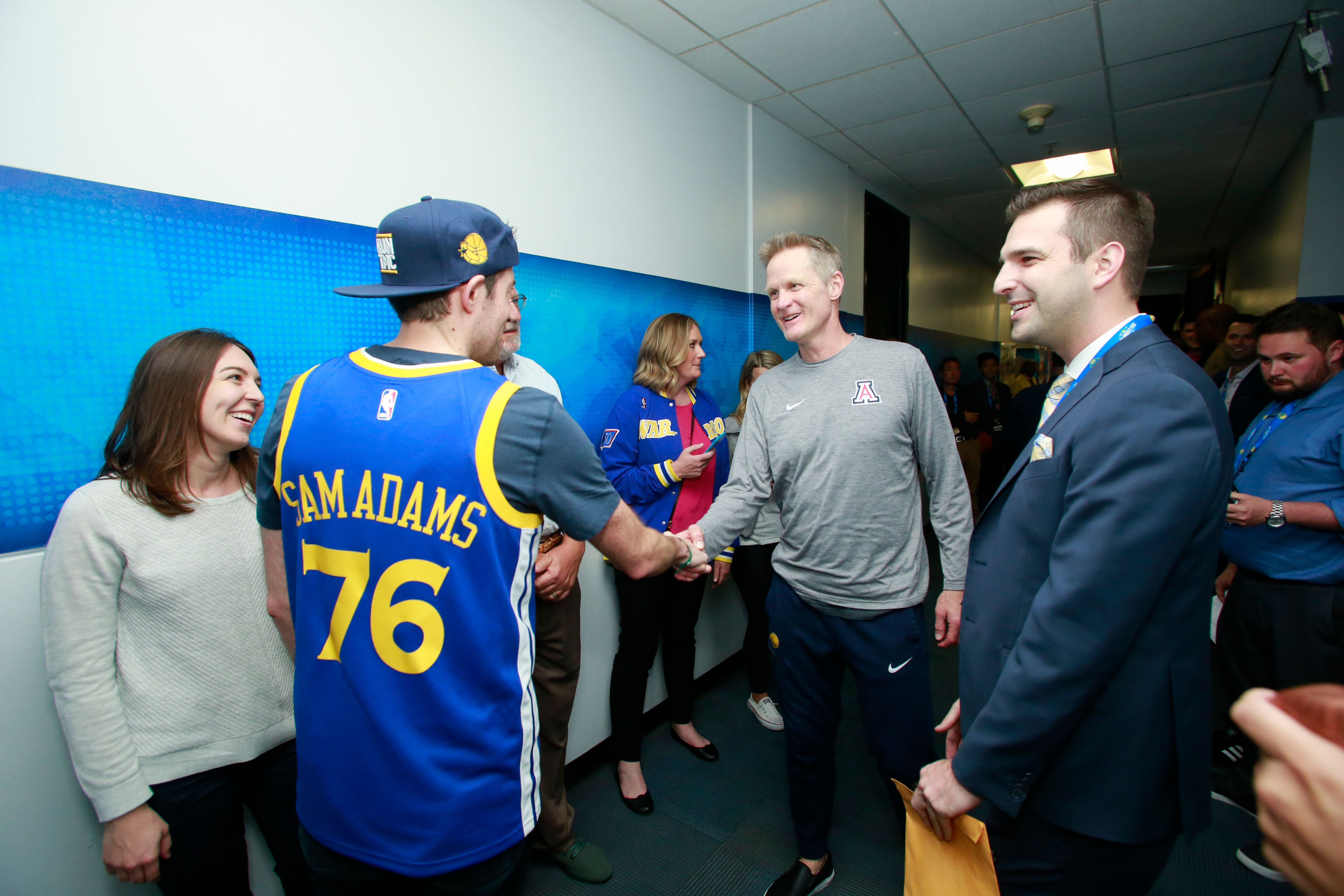 A man in a blue NBA jersey with "SAM ADAMS 76" shakes hands with another